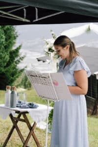 Eine Frau in einem hellblauen Kleid steht an einem dekorativen Notenpult und liest aus einer Mappe. Sie steht im Freien unter einem Baldachin, mit Bergen und Grün im Hintergrund und einem kleinen Tisch mit Flaschen und einem Glas in der Nähe.