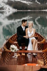 Eine Braut und ein Bräutigam sitzen eng beieinander in einem hölzernen Ruderboot auf einem ruhigen See, umgeben von Bergen. Die Braut trägt ein weißes Kleid und einen Blumenstrauß, der Bräutigam einen schwarzen Anzug, und beide schauen sich liebevoll an.