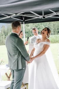 Eine Braut in einem weißen Kleid lacht freudig, während sie während einer Hochzeitszeremonie im Freien mit dem Bräutigam Händchen hält. Im Hintergrund unter einem Baldachin lächeln ein Amtsträger und eine weitere Person.