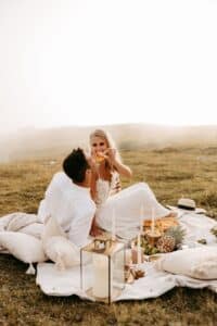 Ein Paar sitzt auf einer Decke im Freien und macht ein Picknick; die Frau füttert den Mann mit einem Stückchen Obst. Sie sind umgeben von Kissen, Kerzen und einer Reihe von Speisen auf einer Wiese bei sanftem, natürlichem Licht.