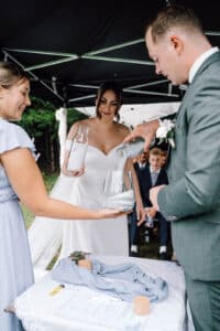 Braut und Bräutigam nehmen an einer Zeremonie teil, bei der sie Sand aus verschiedenen Behältern in ein Gefäß schütten, während eine Frau assistiert und die Gäste im Hintergrund sitzen.