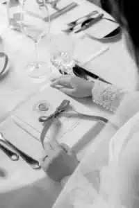 Eine Person in einem langärmeligen Spitzenkleid hält eine Speisekarte mit einer Schleife auf einem elegant gedeckten Tisch mit Gläsern, Besteck und einer gefalteten Serviette. Die Szene wirkt formell und raffiniert.