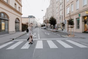 Zwei Personen gehen über einen Zebrastreifen auf einer ruhigen, von Gebäuden und Bäumen gesäumten Straße. Die Straße scheint leer zu sein, es sind weder Autos noch andere Fußgänger in Sicht.
