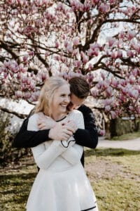 Eine lächelnde Frau in einem weißen Kleid steht vor einem blühenden Magnolienbaum, während ein Mann sie von hinten umarmt. Beide wirken glücklich und verspielt an einem sonnigen Tag im Freien.