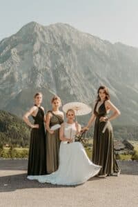 Vier Frauen in formellen Kleidern posieren im Freien vor einem hohen Berg. Die Braut in Weiß sitzt mit einem Sonnenschirm in der Hand, während die drei Brautjungfern in olivgrünen Kleidern zu beiden Seiten stehen, mit einer grünen Kulisse im Hintergrund.