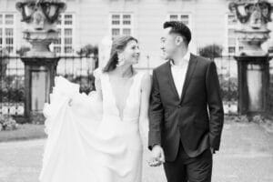 Eine Braut und ein Bräutigam gehen Hand in Hand im Freien und lächeln sich an. Die Braut trägt ein fließendes weißes Kleid und einen Schleier; der Bräutigam trägt einen dunklen Anzug. Im Hintergrund sind verschnörkelte Gebäude und schmiedeeiserne Zäune zu sehen.
