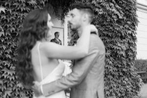 Ein Schwarz-Weiß-Foto zeigt ein Paar, das sich umarmt und im Vordergrund unscharf ist, während ein älterer Mann hinter ihnen, eingerahmt von grünen Ranken, scharf abgebildet ist.