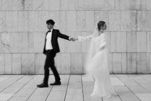 Eine Braut in einem langen weißen Kleid und Schleier hält die Hand eines Bräutigams im Anzug, der sie nach vorne führt; beide bewegen sich vor einem hellen Hintergrund aus Steinfliesen an der Wand.