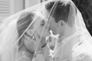 Eine Braut und ein Bräutigam küssen sich romantisch unter einem hauchdünnen Schleier und halten sich gegenseitig das Gesicht. Das Foto ist in Schwarz-Weiß gehalten und unterstreicht den zarten und intimen Moment an ihrem Hochzeitstag.