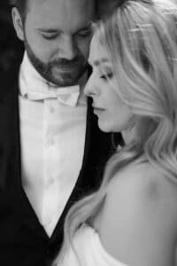 Ein Schwarz-Weiß-Foto eines Mannes im Smoking und mit Fliege, der dicht neben einer Frau mit langem, gewelltem Haar und schulterfreiem Kleid steht, beide wirken gelassen und vertraut.