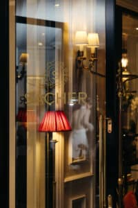 Eine Glastür mit dem goldenen Sacher-Logo führt in einen eleganten Innenraum mit Wandlampen mit weißen und roten Schirmen und kunstvollen Verzierungen, die sich im Glas spiegeln.