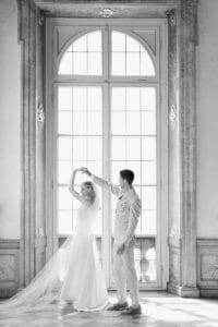 Eine Braut und ein Bräutigam tanzen zusammen vor einem hohen, eleganten Fenster mit fließenden Vorhängen. Die Braut trägt ein langes, weißes Kleid und einen Schleier, der Bräutigam einen hellen Anzug. Sonnenlicht strömt herein und schafft eine romantische Atmosphäre.