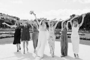 Eine fröhliche Braut und fünf Brautjungfern gehen mit erhobenen Armen, lächelnd und feiernd in einem sonnigen Garten. Die Gruppe trägt verschiedene Kleider und im Hintergrund sind gepflegte Blumenbeete und Bäume zu sehen.