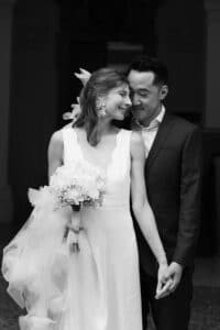Eine Braut in einem weißen Kleid mit einem Blumenstrauß in der Hand lächelt und lehnt sich an einen Bräutigam im Anzug. Sie stehen dicht beieinander, halten sich an den Händen und teilen einen zärtlichen, freudigen Moment. Das Foto ist in schwarz-weiß gehalten.