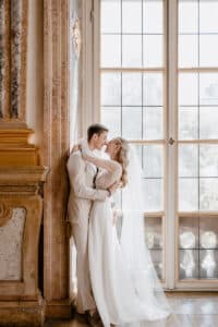 Eine Braut und ein Bräutigam stehen dicht beieinander, umarmen sich und lächeln sich durch große, sonnenbeschienene Fenster in einem eleganten, verzierten Raum mit Marmor- und Goldakzenten an. Die Braut trägt ein weißes Kleid und einen Schleier; der Bräutigam trägt einen hellen Anzug.