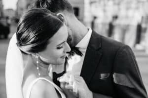 Ein Schwarz-Weiß-Foto von einer Braut und einem Bräutigam. Die Braut, die einen Schleier und baumelnde Ohrringe trägt, lächelt sanft, während der Bräutigam sanft ihre Hand hält und sich dicht an sie lehnt, was einen intimen, romantischen Moment schafft.