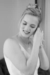 Schwarz-Weiß-Foto einer lächelnden Frau in einem ärmellosen Kleid, die einen Ohrring befestigt. Sie hat ihr helles Haar hochgestylt und trägt eine Halskette. Der Hintergrund ist sanft verschwommen.