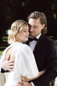 Eine Braut in einem weißen Kleid mit Puffärmeln steht mit geschlossenen Augen da, lächelt sanft und wird von einem Bräutigam im schwarzen Anzug und mit Fliege umarmt, der sie liebevoll anschaut. Sie befinden sich im Freien mit Grünzeug im Hintergrund.