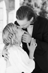 Ein Schwarz-Weiß-Foto eines Paares in formeller Kleidung, das sich einen romantischen Kuss gibt. Der Mann umarmt die Frau sanft, während sie ihren Kopf nach oben neigt und ihre Hand auf seiner Brust ruht. Sie scheinen im Freien zu sein.