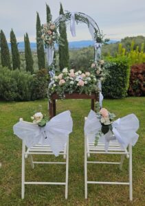 Zwei weiße Stühle, die mit großen weißen Schleifen und Blumen geschmückt sind, stehen vor einem Holztisch, der mit Blumenarrangements geschmückt ist. Hinter dem Tisch steht ein blumengeschmückter Bogen auf einer grasbewachsenen Wiese, auf der im Hintergrund Bäume und Hügel zu sehen sind.