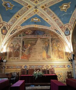 Eine reich verzierte Gewölbedecke und Wände mit komplizierten Fresken und Mustern, die religiöse Figuren darstellen. Darunter befindet sich ein Tisch mit einem Blumengesteck und mehreren Stühlen in einem historischen, reich verzierten Raum.