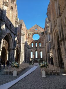 Die Hochzeitszeremonie findet in einer alten Steinabtei ohne Dach statt, mit hohen Bögen, Blumenarrangements und Stuhlreihen vor einem runden Fenster unter einem strahlend blauen Himmel.