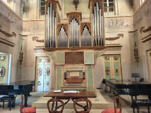 Eine große Pfeifenorgel mit kunstvollem Holzwerk steht in der Mitte einer Bühne in einem eleganten Saal, flankiert von zwei schwarzen Flügeln. Im Vordergrund stehen verzierte Stühle und ein Holztisch mit Papieren und einer Rose.