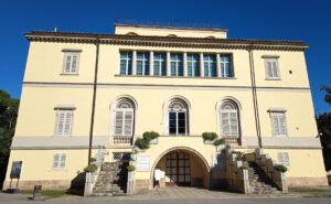 Ein großes, gelbes, dreistöckiges Gebäude mit weißen Fensterläden, Rundbogenfenstern und zwei Treppen, die zu einem zentralen Eingang führen. Topfpflanzen säumen die Treppe, und der Himmel ist klar und blau.