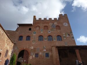 Ein mittelalterliches Backsteingebäude mit gewölbten Fenstern, verschiedenen bunten Wappen an der Fassade, einer Uhr oben rechts und einem blauen Himmel mit Wolken im Hintergrund. Unten stehen Menschen in der Nähe des Eingangs.