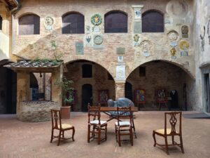 Ein Innenhof mit Backsteinmauern, die mit Wappen und Plaketten verziert sind, mit drei Holzstühlen und einem Tisch in der Mitte, einem steinernen Brunnen auf der linken Seite und Kunstwerken unter Steinbögen im Hintergrund.