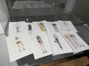 Neun aquarellierte Modeillustrationen sind auf einem reflektierenden Tisch ausgestellt. Sie zeigen unterschiedliche Outfits und Stile, die von verschiedenen Figuren, sowohl Einzelpersonen als auch Paaren, in einer Vielzahl von Posen getragen werden.