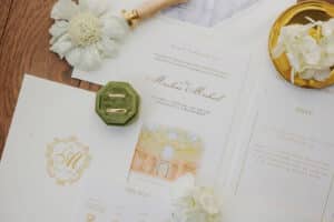 Elegantes Hochzeitspapeterie-Set mit goldenen und weißen Einladungen, einer Antwortkarte, einer samtgrünen Ringschachtel mit zwei goldenen Ringen, weißen Blumen und einem goldenen Wachssiegelstempel, alles auf einer hellen Oberfläche angeordnet.