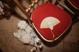 Ein rot gepolsterter Goldstuhl mit einem illustrierten Fächer und einer Quaste auf dem Sitz, neben weißen Blumen und goldenen Kerzen auf einem Marmorboden.