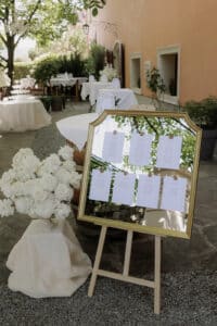 Ein goldgerahmter Spiegel auf einer Staffelei zeigt ein Blatt Papier mit Text, wahrscheinlich ein Sitzplan für eine Hochzeit. Er steht im Freien neben einem großen Strauß weißer Blumen und einem pfirsichfarbenen Gebäude, mit Tischen im Hintergrund.