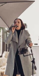 Eine Frau in einem grauen Mantel singt im Freien in ein Mikrofon und hält einen Ständer mit einem Tablet in der Hand. Im Hintergrund sind ein offener Regenschirm und einige unscharfe Objekte zu sehen.