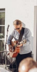 Ein Mann mit Sonnenbrille und hellblauem Hemd spielt vor einem Mikrofon auf einer Akustikgitarre. Er steht im Freien neben einer weißen Wand im hellen Sonnenlicht.