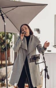 Eine Frau in einem grauen Mantel singt im Freien in ein Mikrofon, das unter einem großen Regenschirm neben einem Notenständer mit einem Tablet steht.