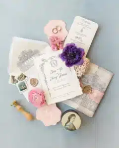 Flaches Layout von eleganten Hochzeitspapieren, einschließlich Einladungen mit architektonischen Skizzen, Vintage-Stempeln, Wachssiegeln, Ringen, rosa und lila Blumen und einer kleinen Dose mit einem klassischen Porträt auf einem weichen blauen Hintergrund.