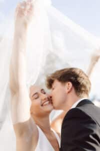 Eine fröhliche Braut lächelt mit geschlossenen Augen, während ihr Bräutigam sie auf die Wange küsst. Die Braut lüftet den durchsichtigen Schleier über ihnen, und beide wirken glücklich an einem hellen, sonnigen Tag.