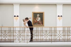 Braut und Bräutigam küssen sich auf einem eleganten Balkon mit weißen Wänden, goldenem Geländer, schwarz-goldenen Wandleuchten und einem gerahmten historischen Porträt im Hintergrund.