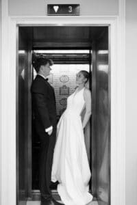 Eine Braut und ein Bräutigam stehen sich in einem Aufzug gegenüber, die Braut in einem weißen Kleid und der Bräutigam in einem schwarzen Anzug, unter einer Anzeige mit der Zahl 3. Die Szene ist in schwarz-weiß gehalten.