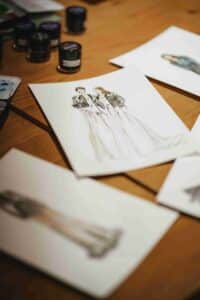 Nahaufnahme von Modeskizzen auf einem Holztisch, mit zwei Figuren in langen Kleidern und Jacken als zentraler Zeichnung, um die herum Kunstzubehör und andere Skizzen verstreut sind.