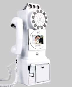 Ein weißes altes Münztelefon mit Münzeinwurf und Hörer, auf dessen kleinem Bildschirm ein sich küssendes Paar in Hochzeitskleidung zu sehen ist. Das Telefon ist vor einem schlichten grauen Hintergrund gesetzt.