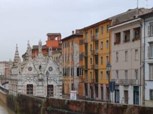 Bunte Gebäude und die gotische Kirche Santa Maria della Spina aus weißem Marmor säumen den Fluss Arno in Pisa, Italien, bei bedecktem Himmel.