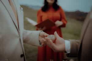 Zwei Personen in Anzügen tauschen während einer Zeremonie im Freien einen kleinen runden Stein aus. Im unscharfen Hintergrund steht eine Person in einem orangefarbenen Kleid, die ein Buch hält.