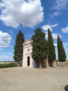 Eine kleine Steinkapelle mit dreieckigem Giebel und rundem Fenster steht zwischen hohen Zypressen unter einem strahlend blauen Himmel mit vereinzelten Wolken, umgeben von offener Landschaft.