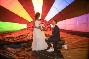 Eine Braut in einem weißen Kleid hält einen Blumenstrauß, während der Bräutigam im Anzug vor ihr in einer bunten, aufgeblasenen Heißluftballonhülle kniet. Leuchtende Regenbogentafeln umgeben das Paar.