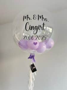 Ein großer durchsichtiger Ballon mit kleinen lila Ballons im Inneren, verziert mit "Mr. & Mrs. Cingirt 21.06.2025" in schwarzer Schrift und einem lila Herz. Eine lila Schleife und weiße Luftschlangen hängen unten mit einem schwarzen Geschenkanhänger.