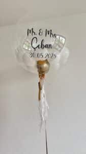 Ein durchsichtiger Ballon mit weißen und goldenen Luftballons im Inneren. Der Text auf dem Ballon lautet "Mr. & Mrs. Çoban 30.05.2025". An der Unterseite hängen weiße und goldene Bänder. Der Ballon steht vor einer schlichten weißen Wand.