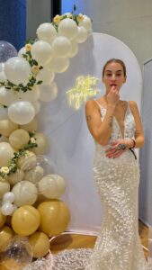 Eine Frau in einem weißen Spitzenhochzeitskleid steht vor einem Ballonbogen mit weißen und goldenen Luftballons und einem Neonschild mit der Aufschrift "Better Together". Sie berührt ihre Lippen und schaut in die Kamera.
